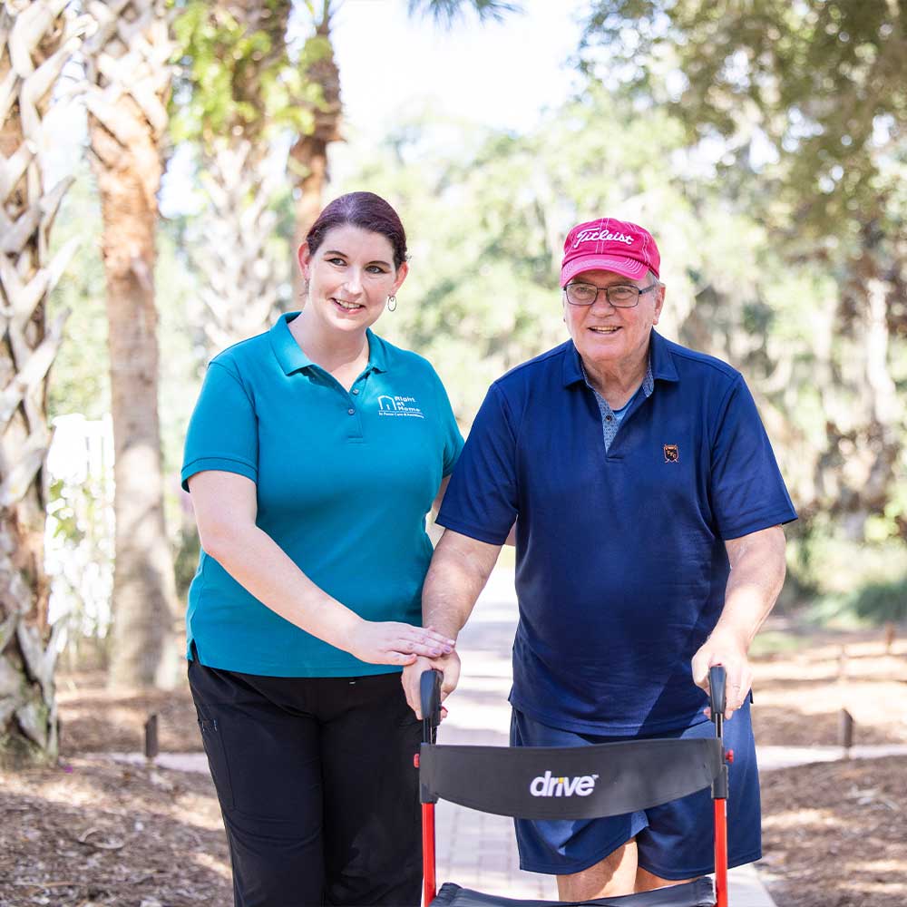 Caregiver helping senior walk along brick pathway between palm trees