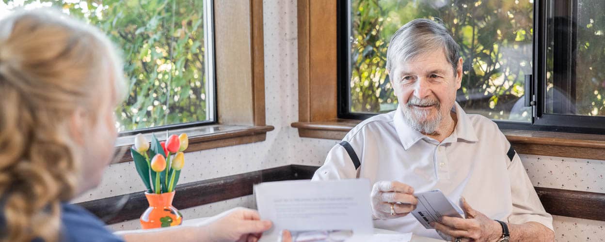 Senior male sitting at a kitchen table with a Right at Home representative discussing the benefits of in home care