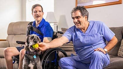 Adult male client with disabilities sitting in a wheelchair next to his male Right at Home caregiver, who is holding a ball in a dog's mouth