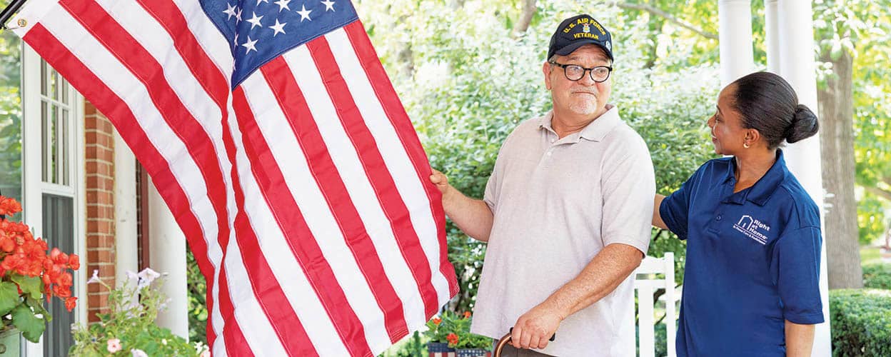 A female Right at Home caregiver is standing next to a senior male veteran client who is hanging up an American flag on his porch. 