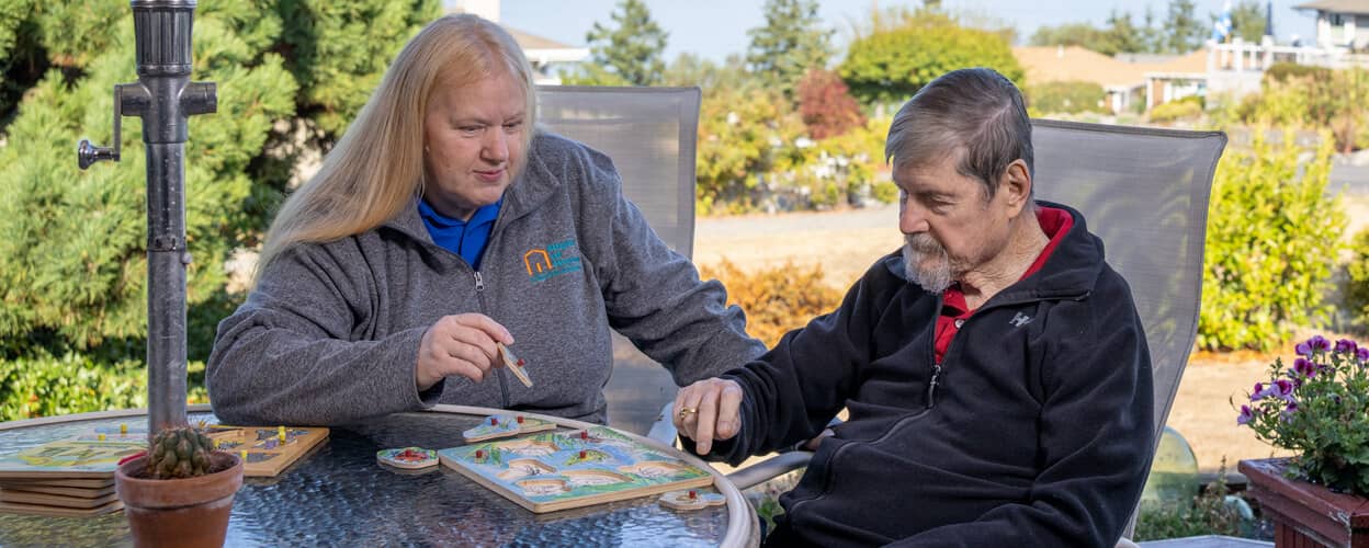 A female Right at Home caregiver is helping a senior male with Dementia solve a puzzle while sitting outside at a patio table