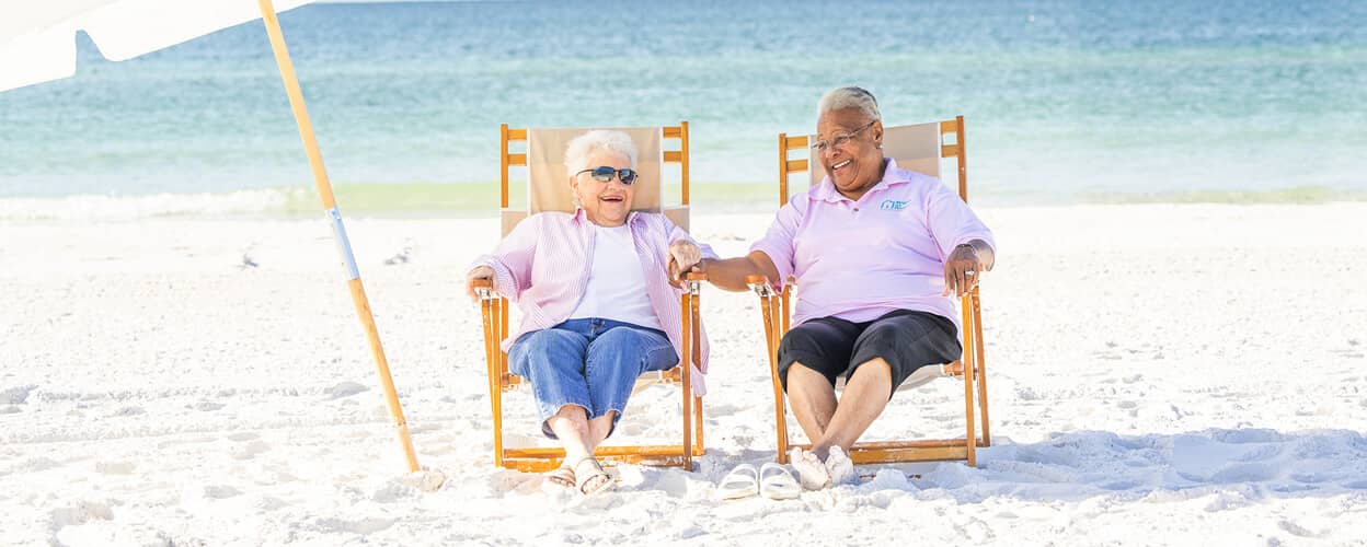 Caregiver and client sitting in lounge chairs under an umbrella on the beach.