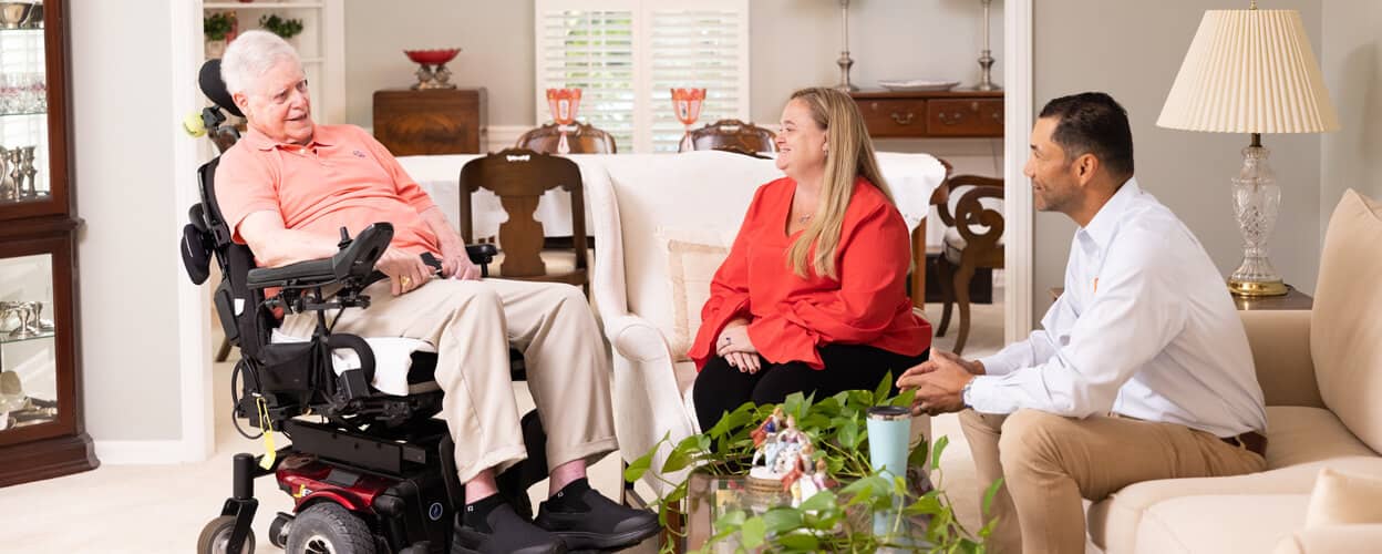 A man and a woman talking to a senior male in a wheelchair in a living room
