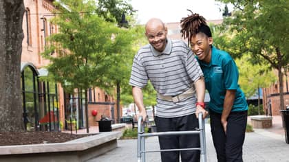 A female Right at Home caregiver helping a man walk down a sidewalk who is using a walker while smiling outside