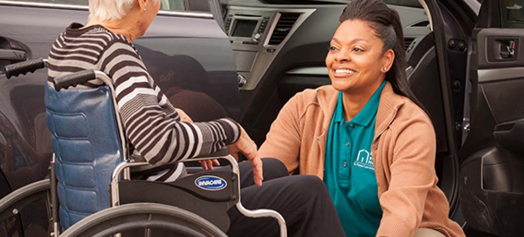 A female caregiver smiles up at her client as she prepares her wheelchair.