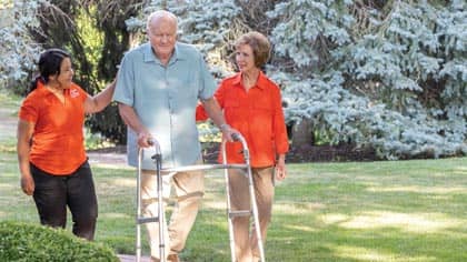 Senior with caregiver and his wife, assisted walking outside.