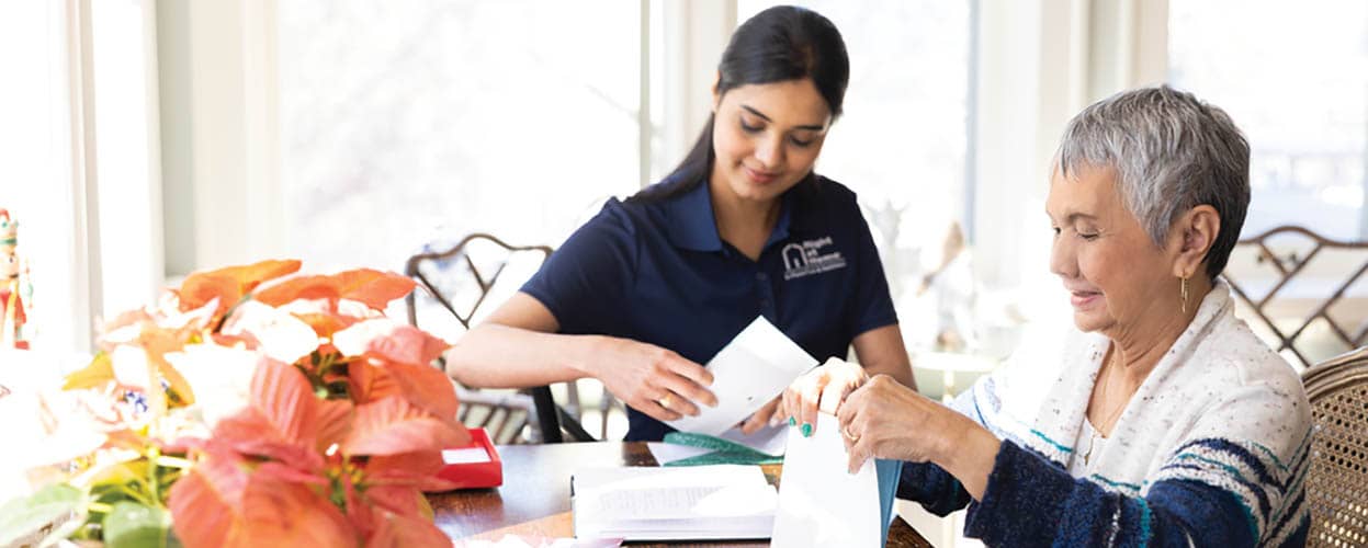 A female Right at Home caregiver helping a senior female stuff envelopes at the kitchen table