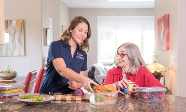 Female caregiver wearing mask serves plate of food to smiling elderly patient.