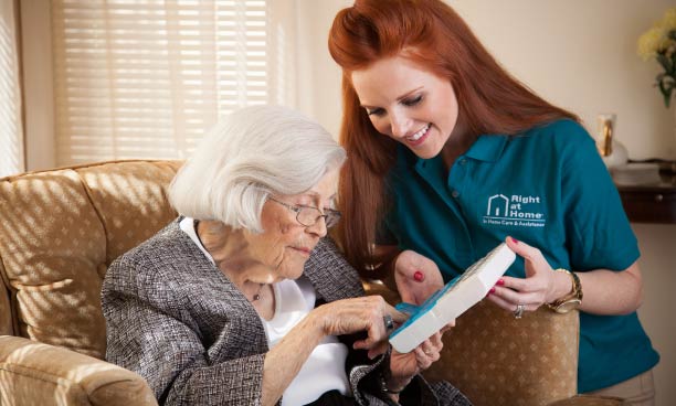 Smiling female caregiver shows an electronic device to elderly female sitting on a couch.