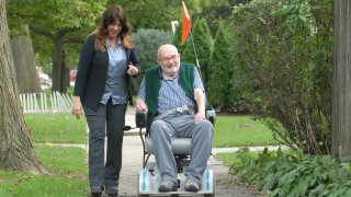 A female caregiver walks on the sidewalk with male senior in a wheelchair. 