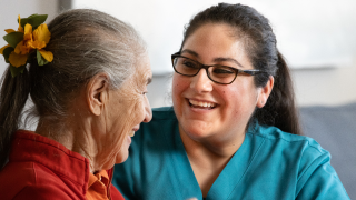 A female caregiver sits across from an elderly female patient, looking at her. They both smile.