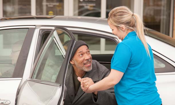 Female caregiver helps elderly patient out of the back seat of a car.